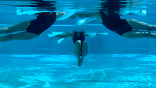 Woman performing a pool pilates side to side exercise, tucking knees and extending legs with a noodle.