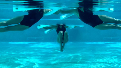 Woman performing a pool pilates side to side exercise, tucking knees and extending legs with a noodle.