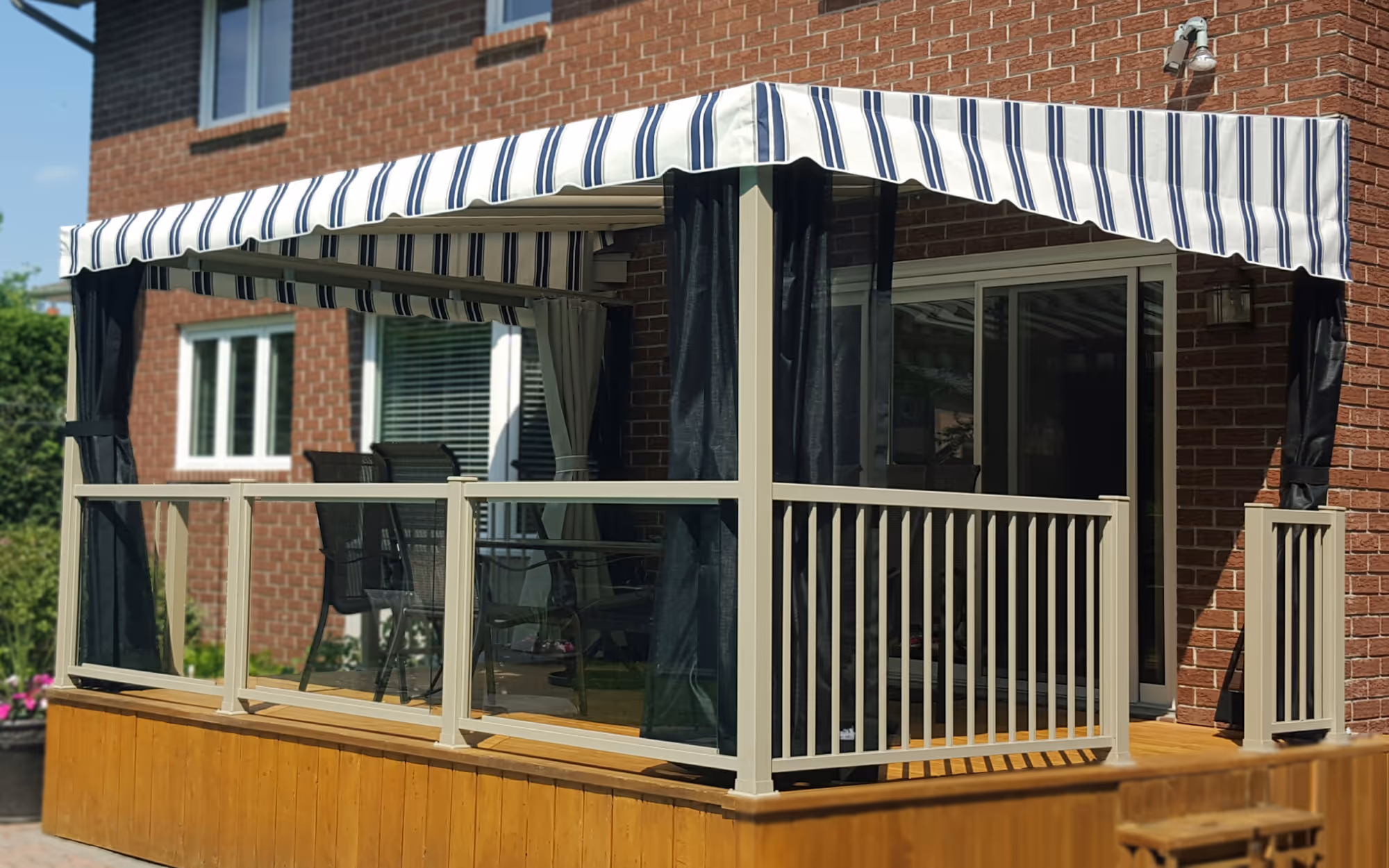 Wooden deck attached to a brick house with tan railings built into a fixed awning with matching tan frame. The awning cover is a striped blue-and-white fabric awning.