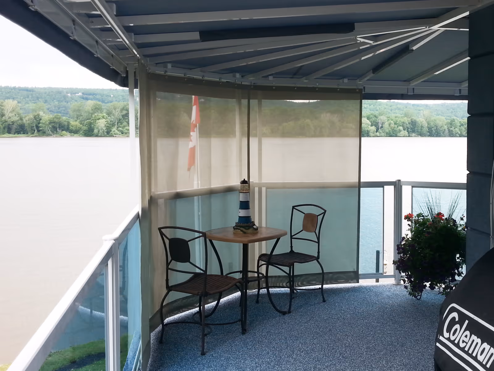 Covered balcony overlooking a river with a seating area for two, sun mesh curtains, and a potted plant.