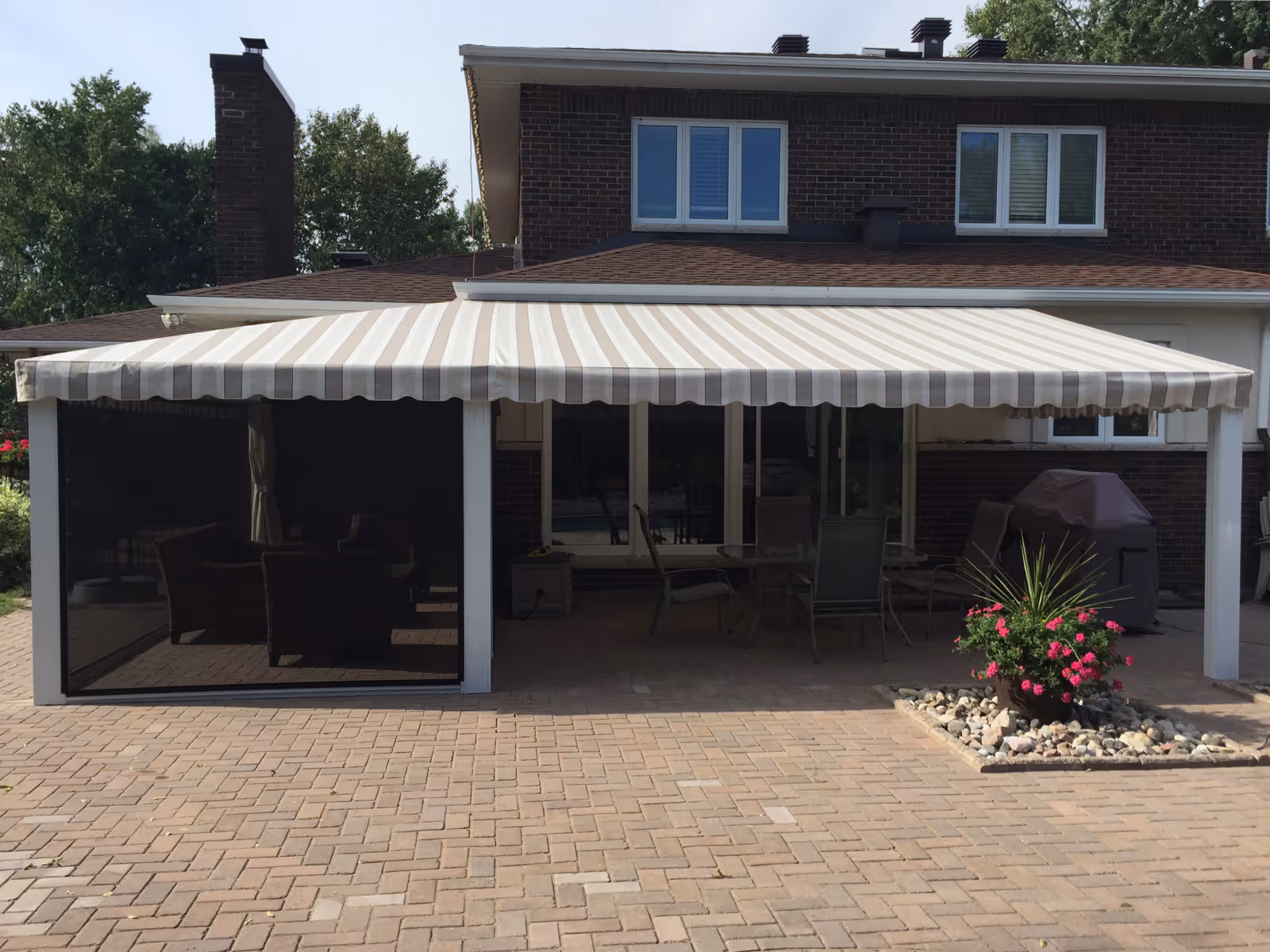 Stone patio area with beige and white striped fixed awning and black tension screen enclosure, furnished with chairs and table, attached to a brick house.