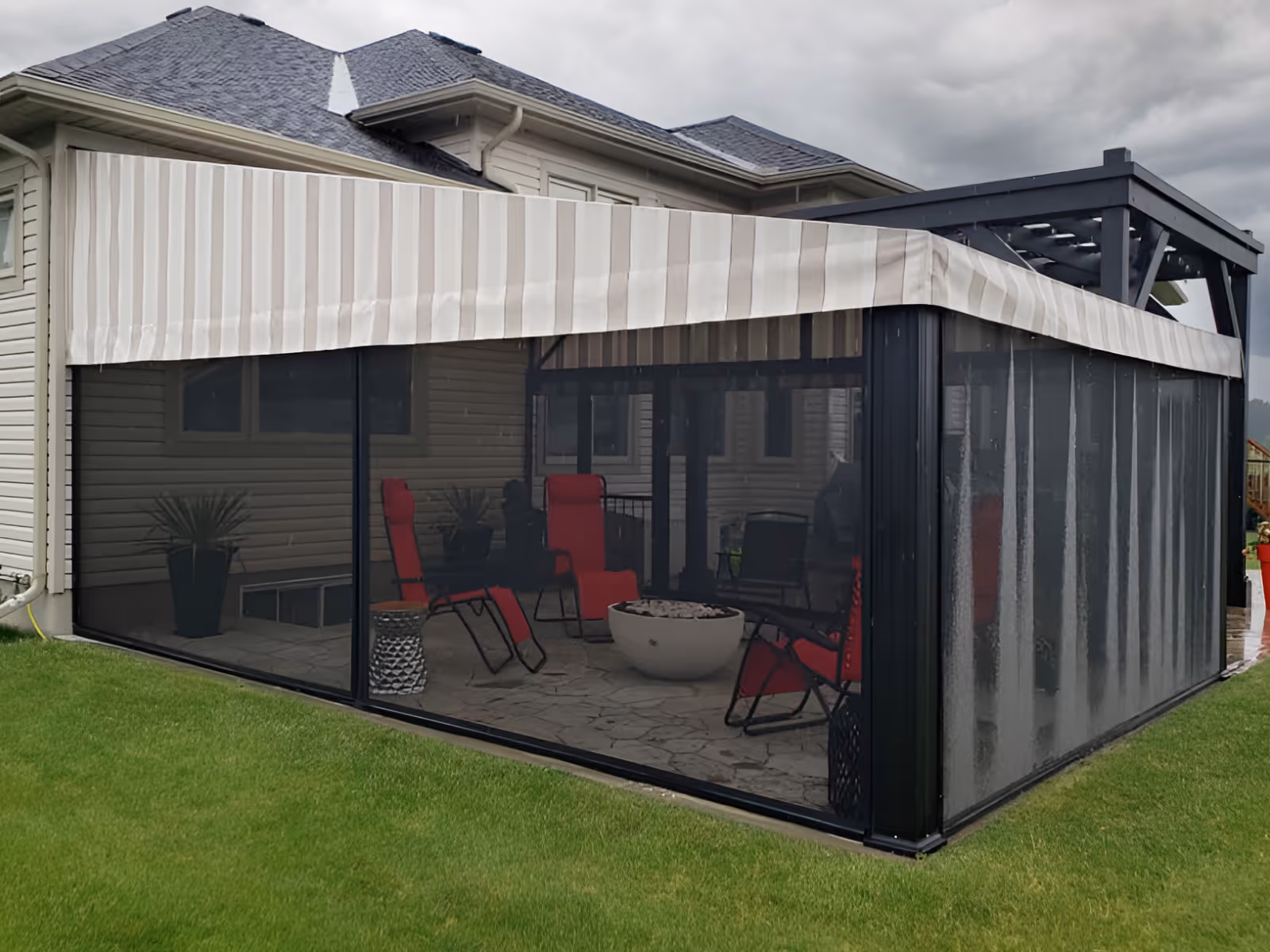 Green lawn surrounds a large stone patio covered by a fixed awning with beige striped fabric.