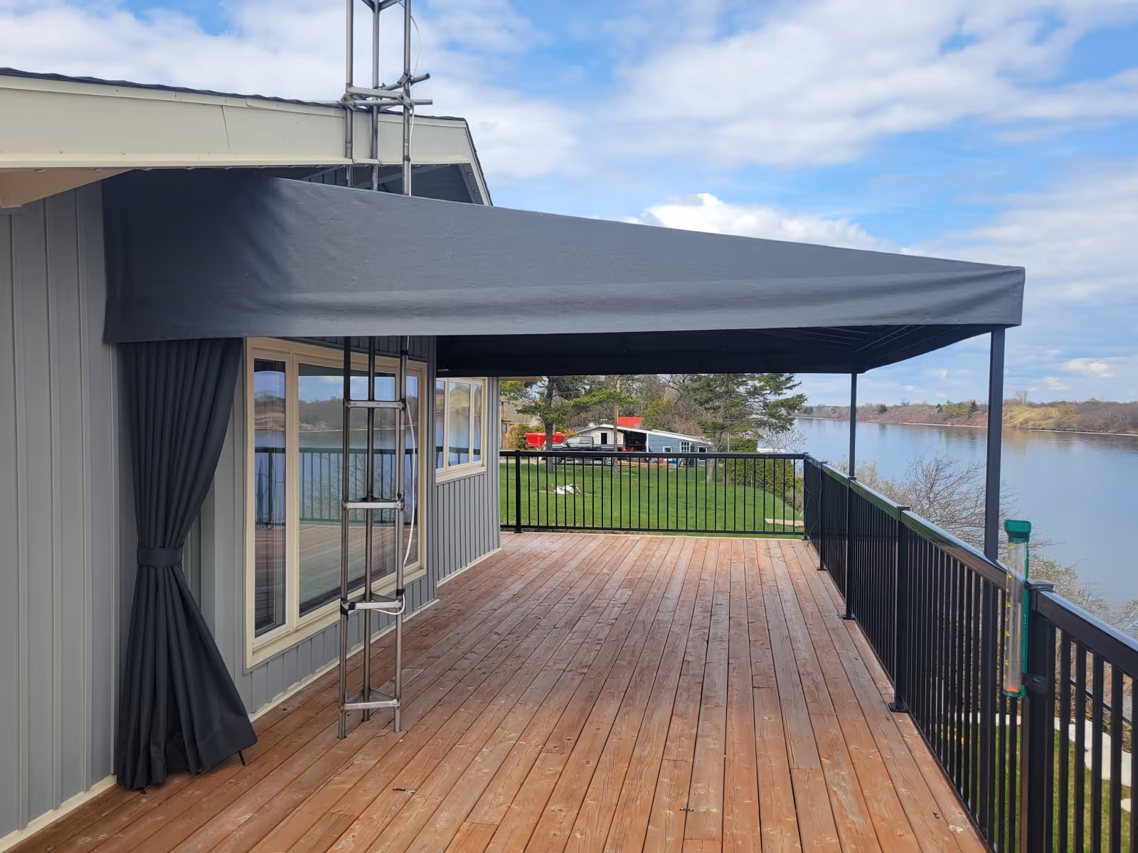 Spacious wooden deck with black metal railing, covered by a large grey awning attached to a grey house overlooking a river.