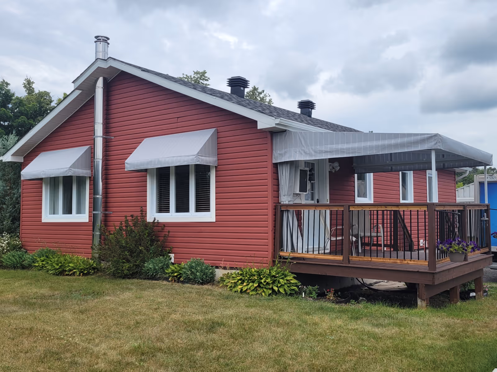Red house with white-trimmed windows featuring matching grey striped awnings over the windows and wood deck.