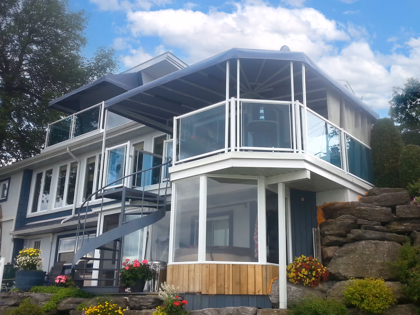Modern two-story house with glass railings on the balcony, spiral staircase, and a covered awning on the upper deck surrounded by rocks and colorful flowers.