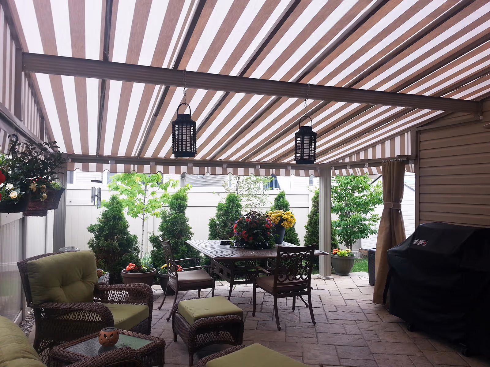 Patio with striped retractable awning, metal dining table with chairs, wicker seating with green cushions, hanging lanterns, and potted plants.