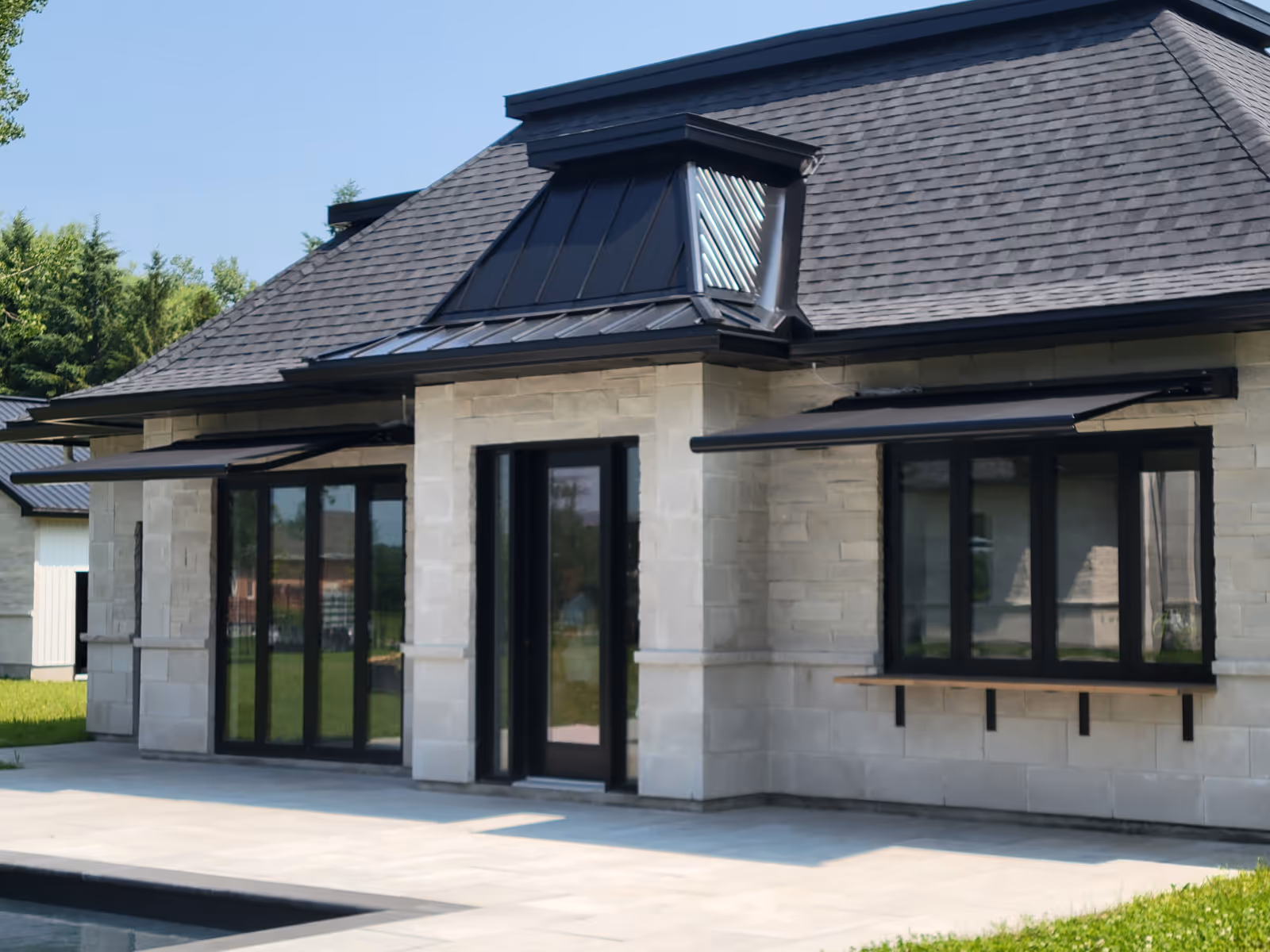 Modern house exterior with beige stone walls, black-framed windows and doors, and two extended black retractable awnings above the windows.