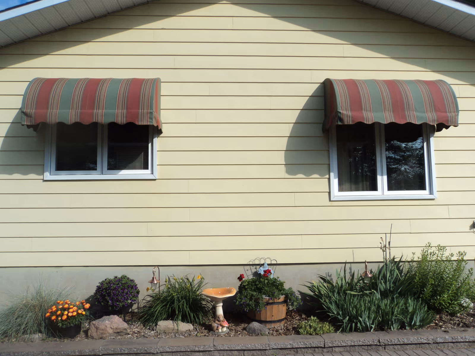 Yellow house exterior with two white-framed windows each covered by red and green striped awnings above a garden bed with various plants and flowers.
