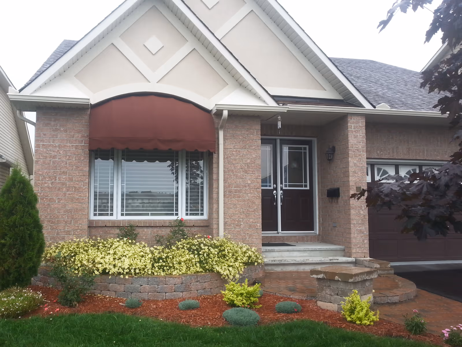 Brick house front with a maroon clamshell awning over a large window, double front doors, and landscaped garden with yellow flowers and shrubs.