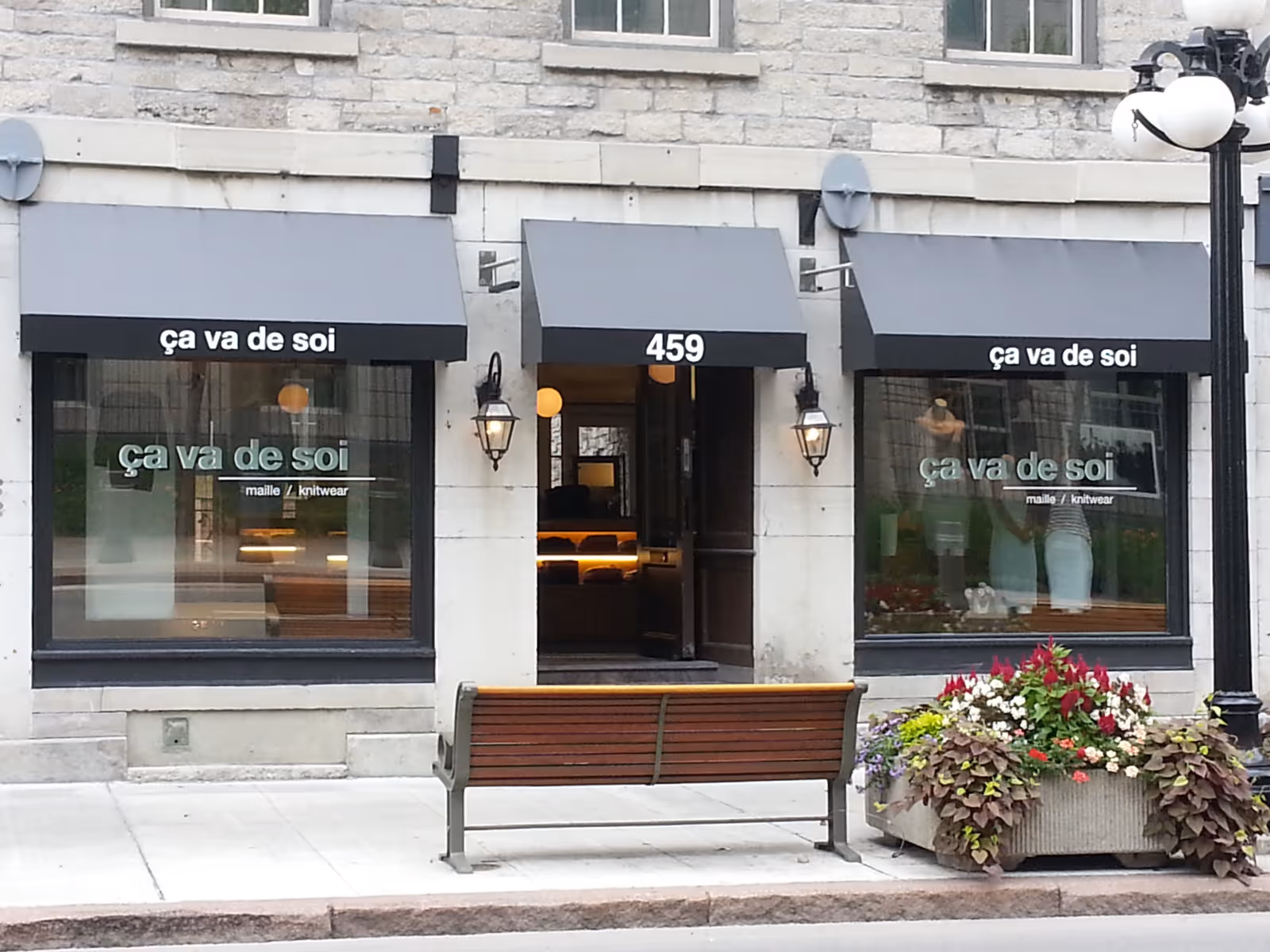 A boutique in an historic stone building with three black awnings displaying the store's name and street number faces an urban sidewalk.