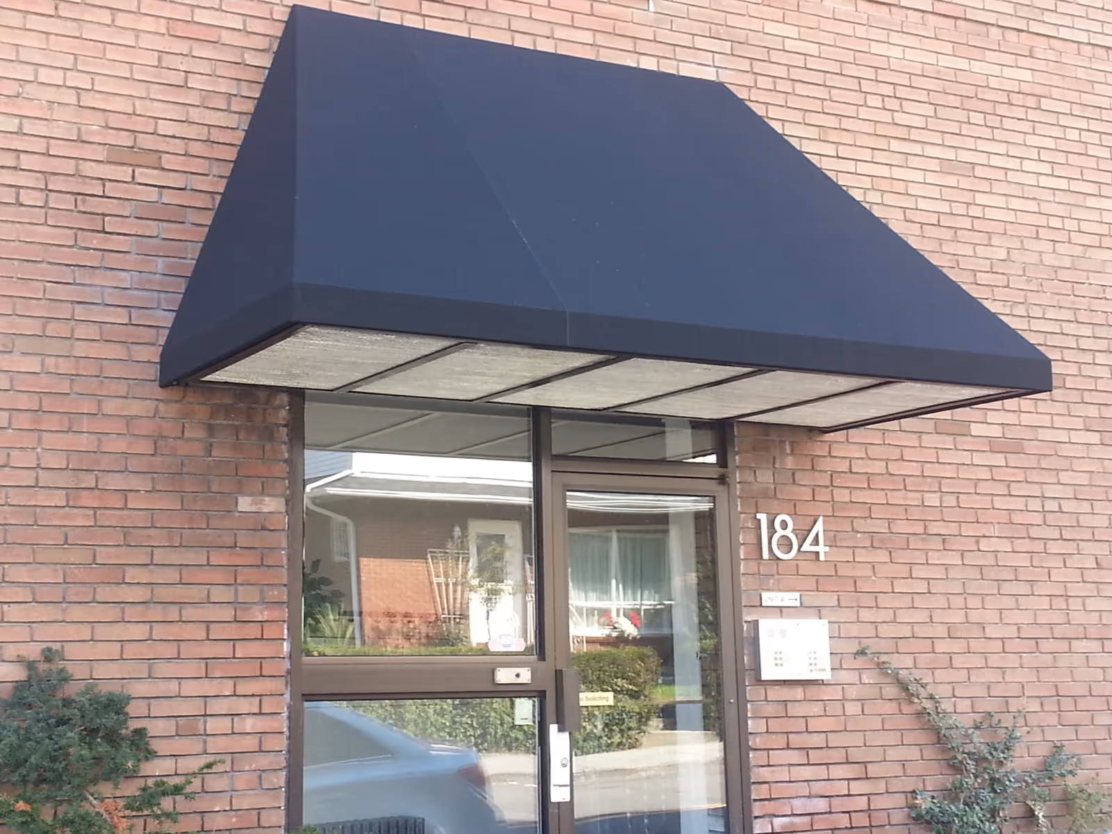 Black trapezoid-shaped awning on a brick building hangs over a glass door.
