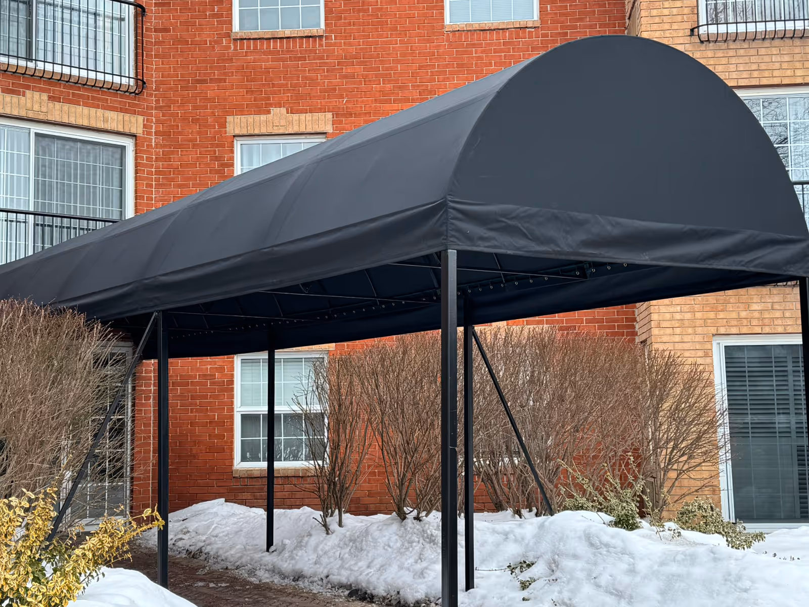 Black arched awning covering a brick walkway entrance beside snow-covered bushes and a red brick building.