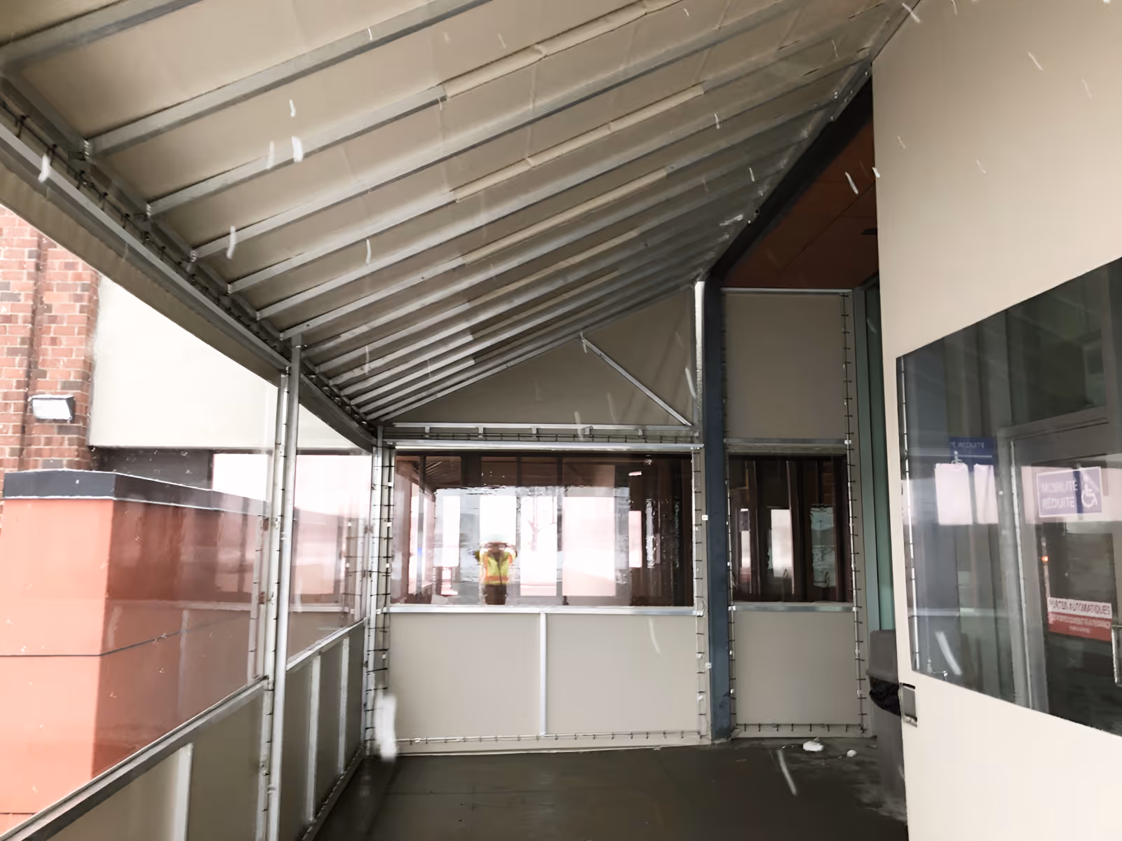 Enclosed walkway with a slanted fabric awning roof and panels with clear viny windows attached to a building entrance.