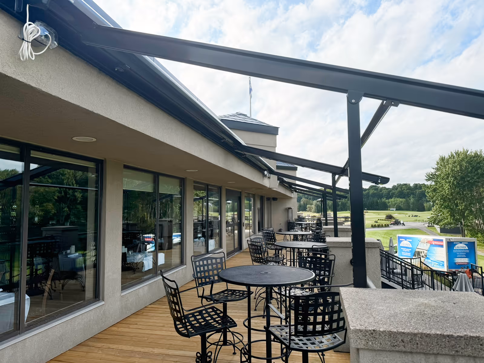 Outdoor restaurant patio with black metal tables and chairs under black pergolino awnings overlooking a green golf course.