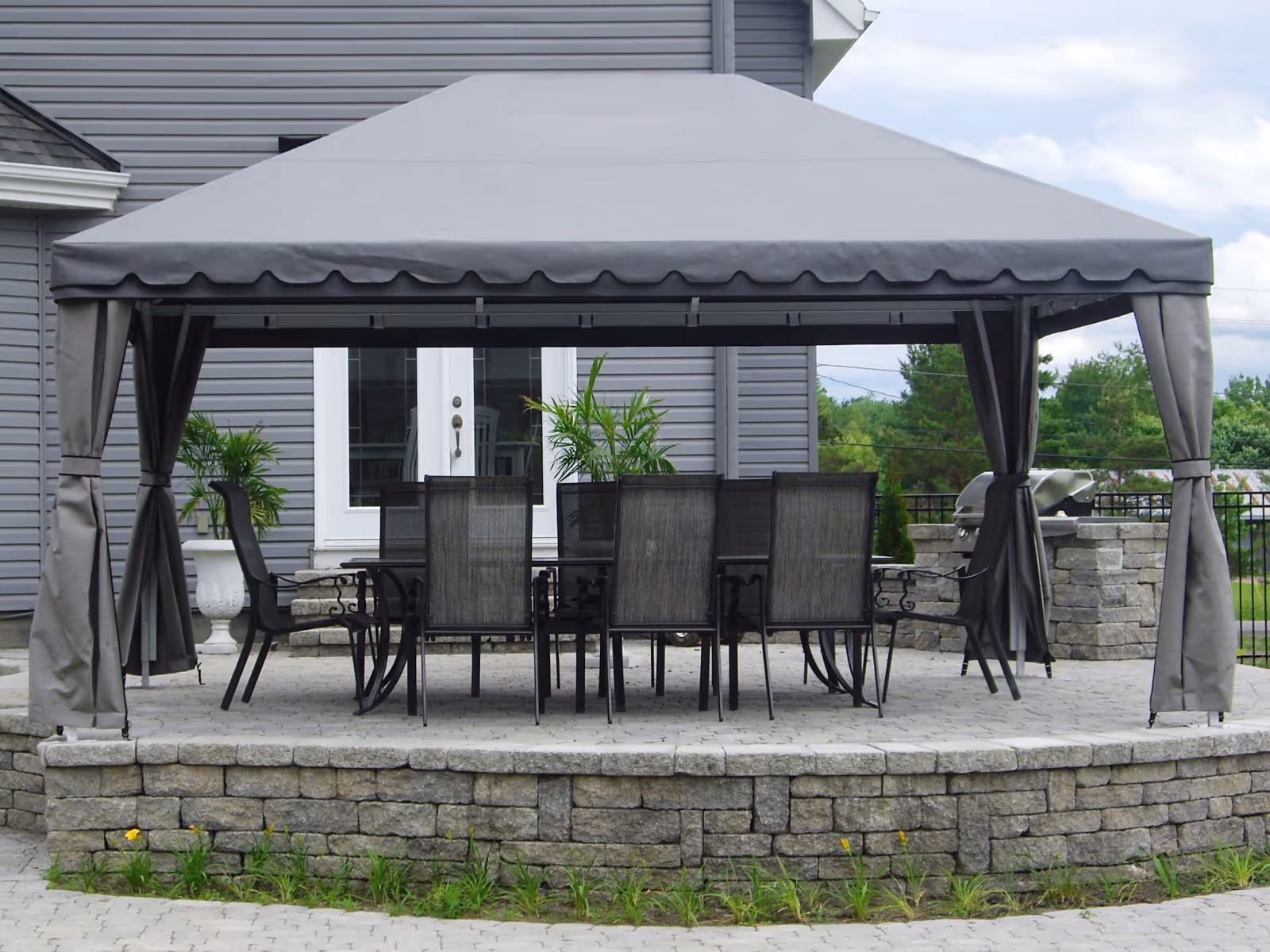 Elevated stone patio next to a grey house with dining table, chairs, and a grey fabric gazebo in a backyard.