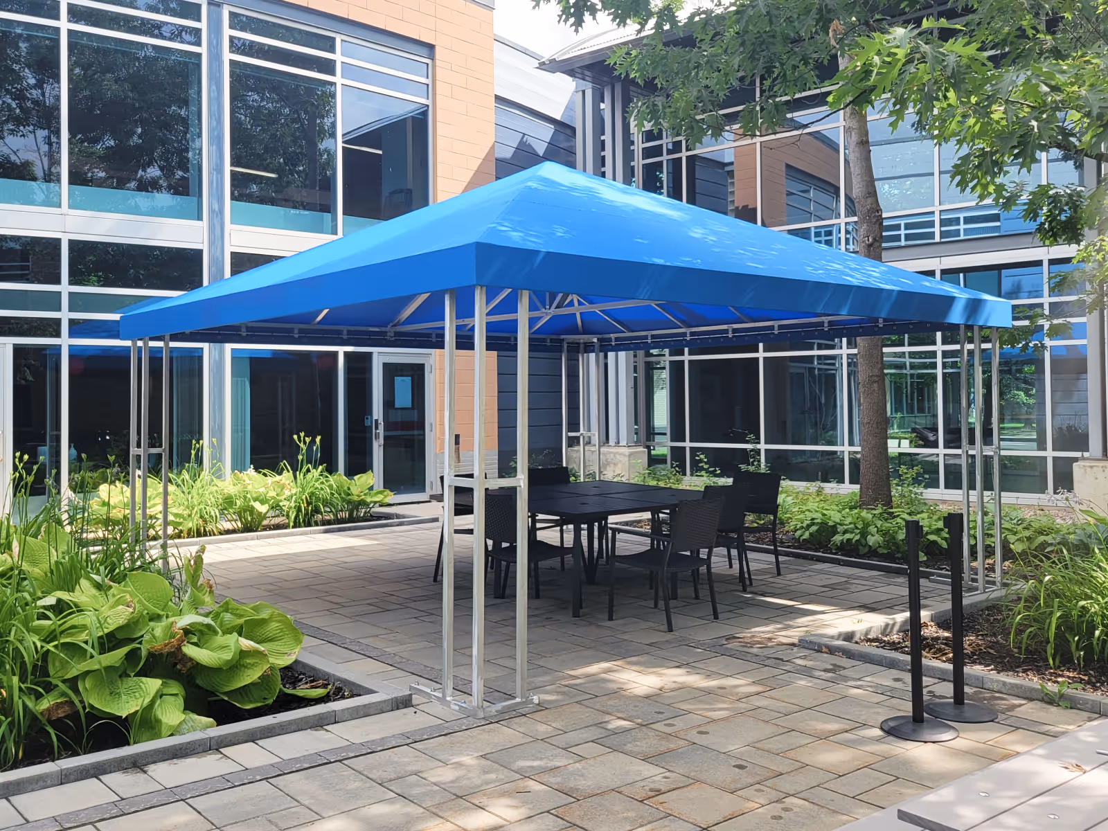 Square gazebo with blue fabric covers a table and chairs in a landscaped courtyard.