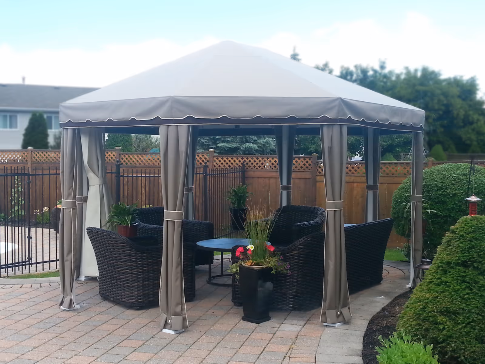 Octagonal gazebo with taupe canopy covers black wicker chairs and a glass-top table on a stone patio.