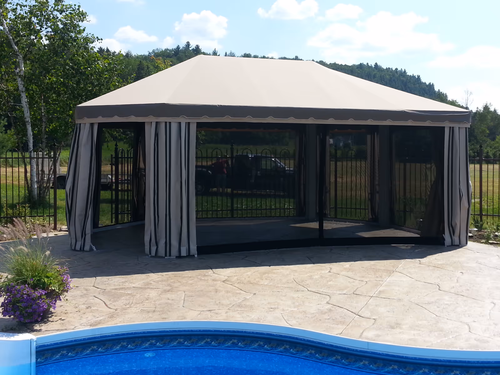 Large beige gazebo on the stone surround of a curvy swimming pool.
