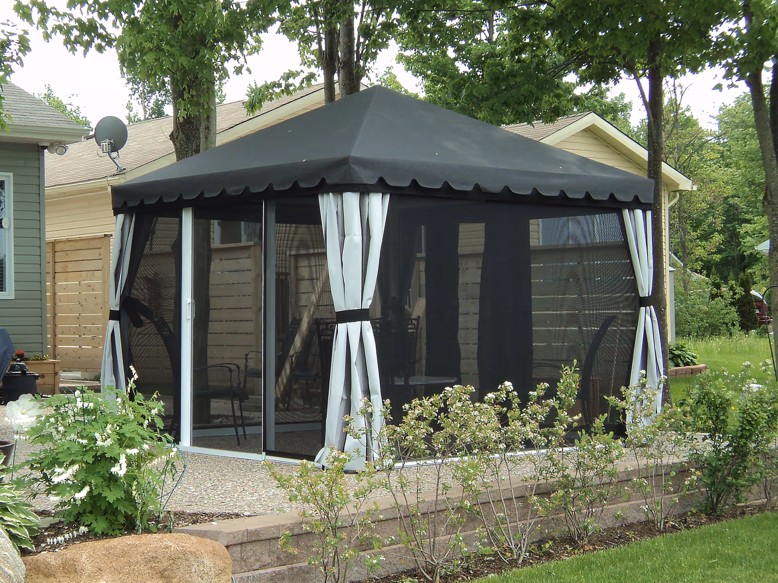 Backyard gazebo with grey canopy, mesh screens, and light grey hide-a-posts, surrounded by garden plants and trees.