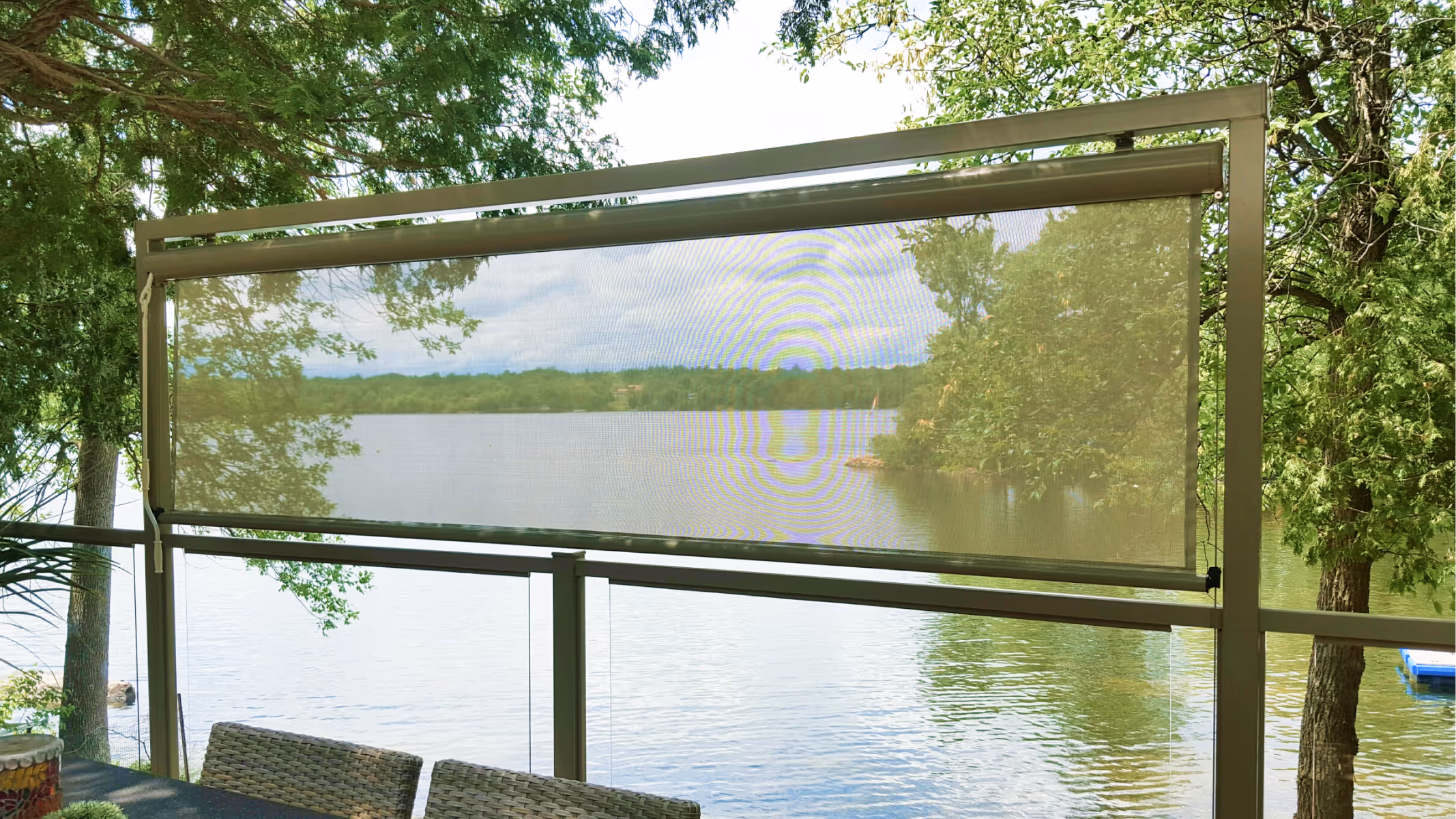 A short exterior roller blind with beige sun mesh hangs over a small section of a glass railing, partially obscuring the view of the water beyond.