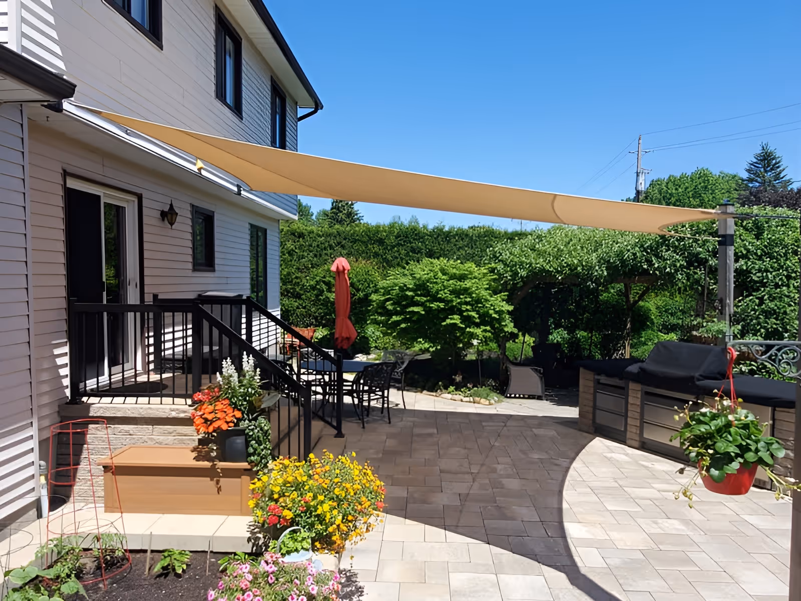 Backyard patio with a beige shade sail, stone pavers, outdoor furniture, and vibrant garden plants.