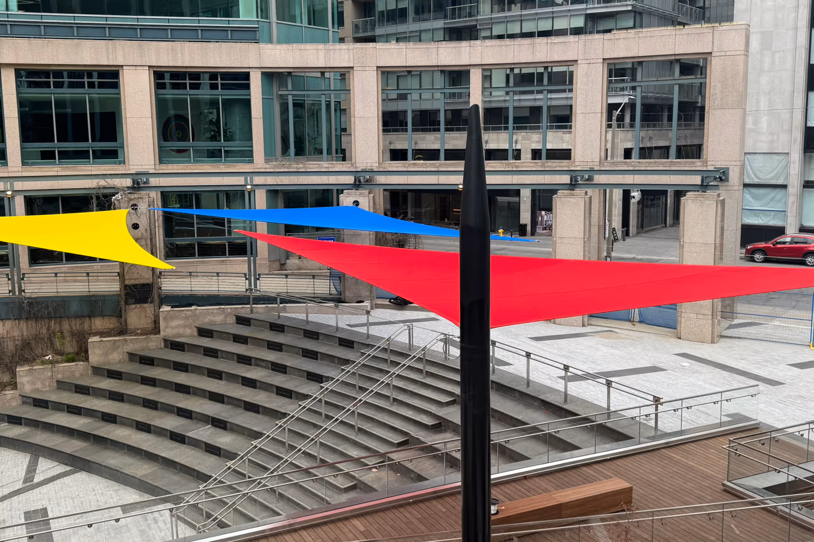 An office building courtyard with tiered concrete seating and three colorful triangular shade sails in yellow, blue, and red suspended above.