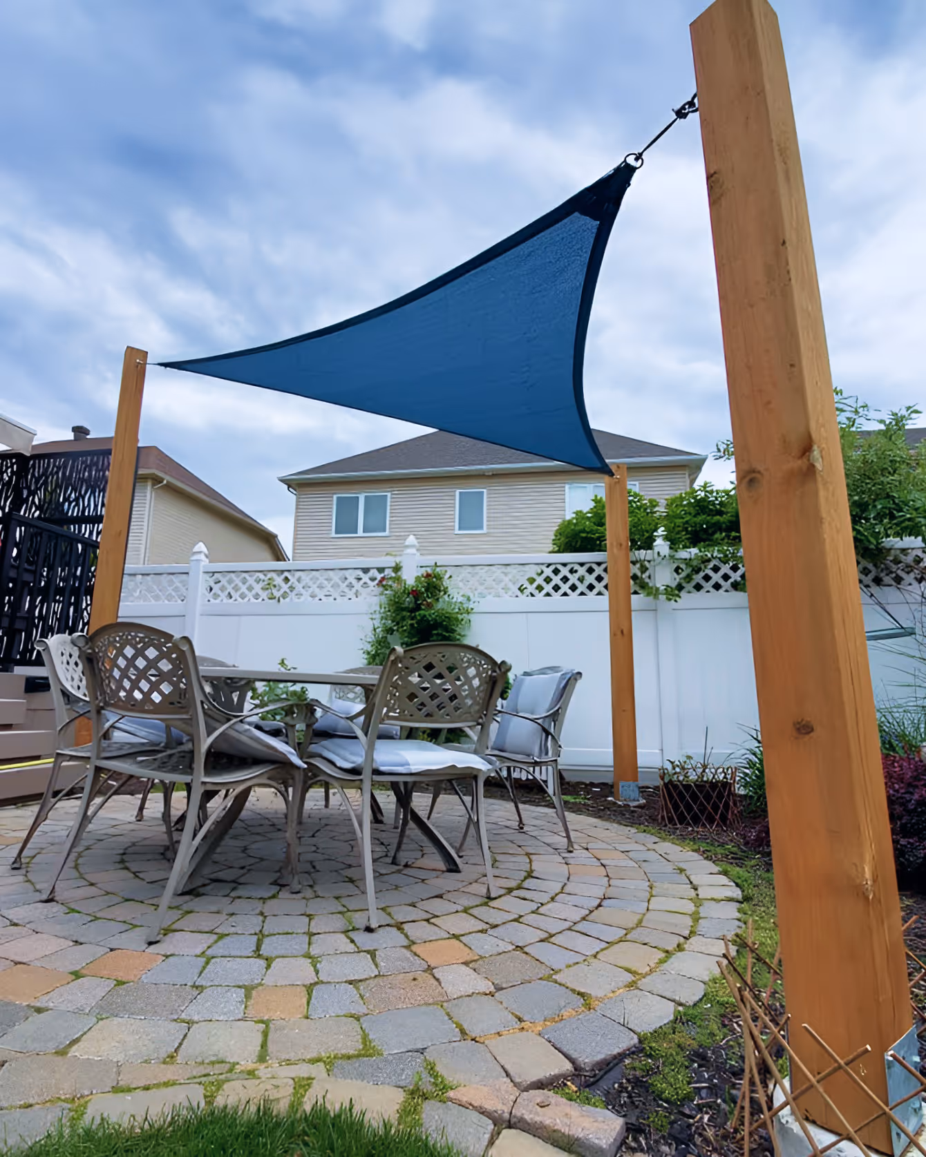 Backyard patio with a round stone paver area, metal chairs with cushions, and a blue triangular shade sail attached to wooden posts.