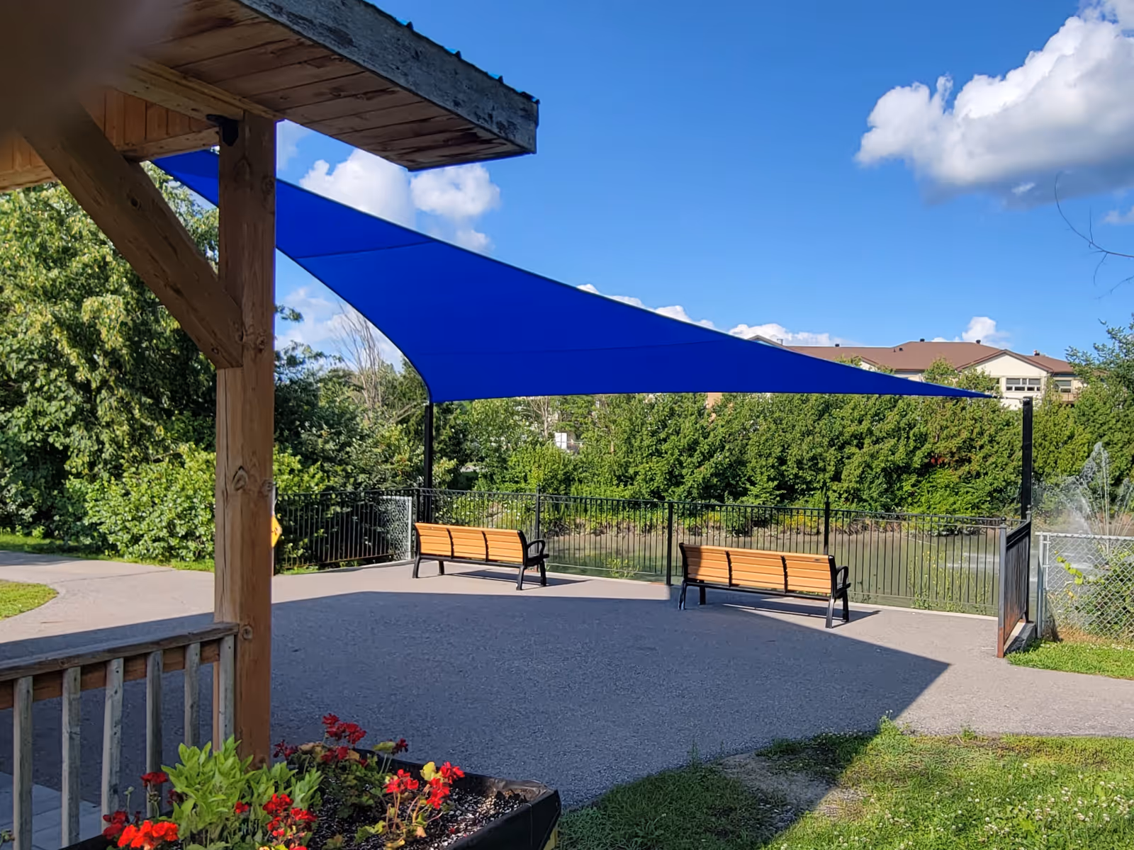 Blue triangular shade sail over two wooden benches at a paved lookout point with greenery and a water fountain in the background.