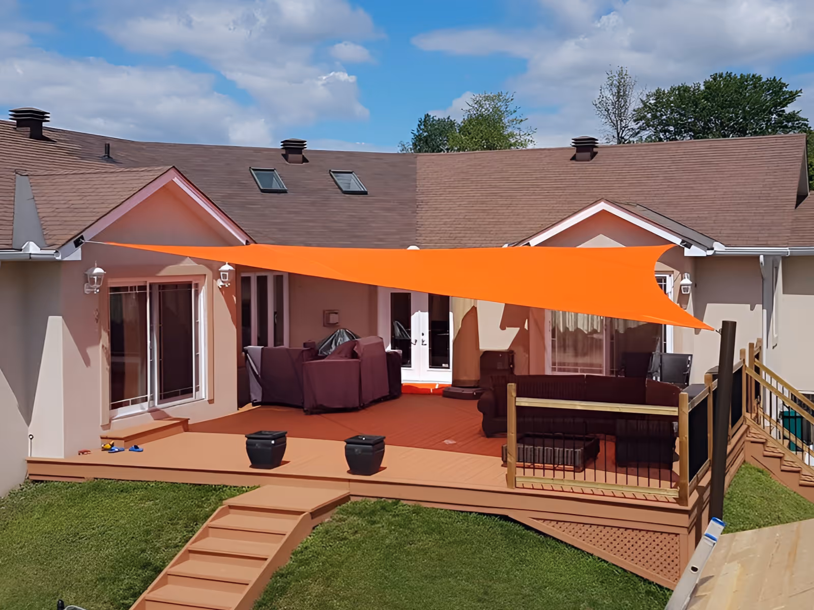 Backyard wooden deck with patio furniture shaded by a bright orange triangular shade sail attached to a beige house.