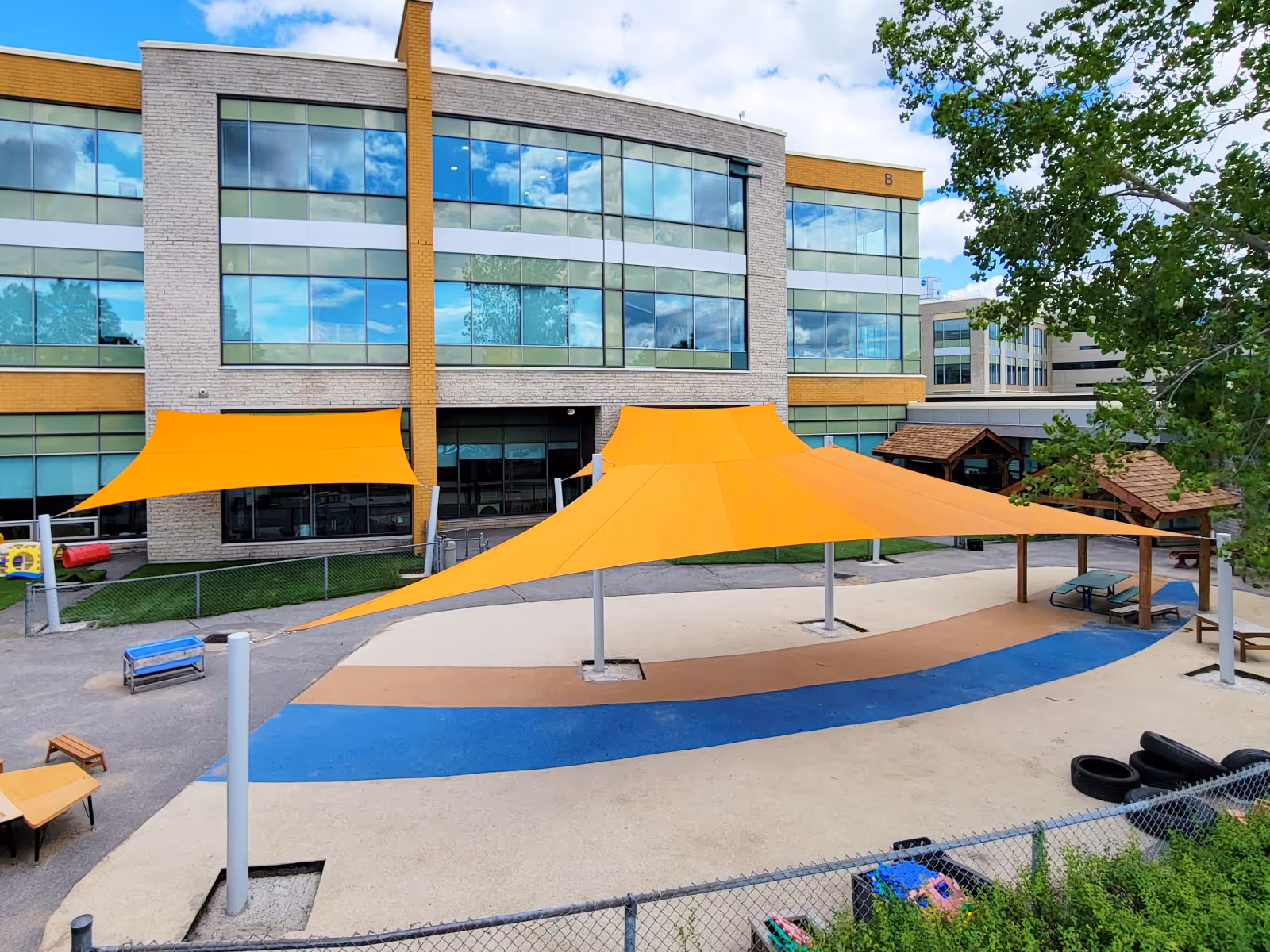 Outdoor recreation area with large orange shade sails over a blue and beige paved surface next to a modern building with reflective windows.