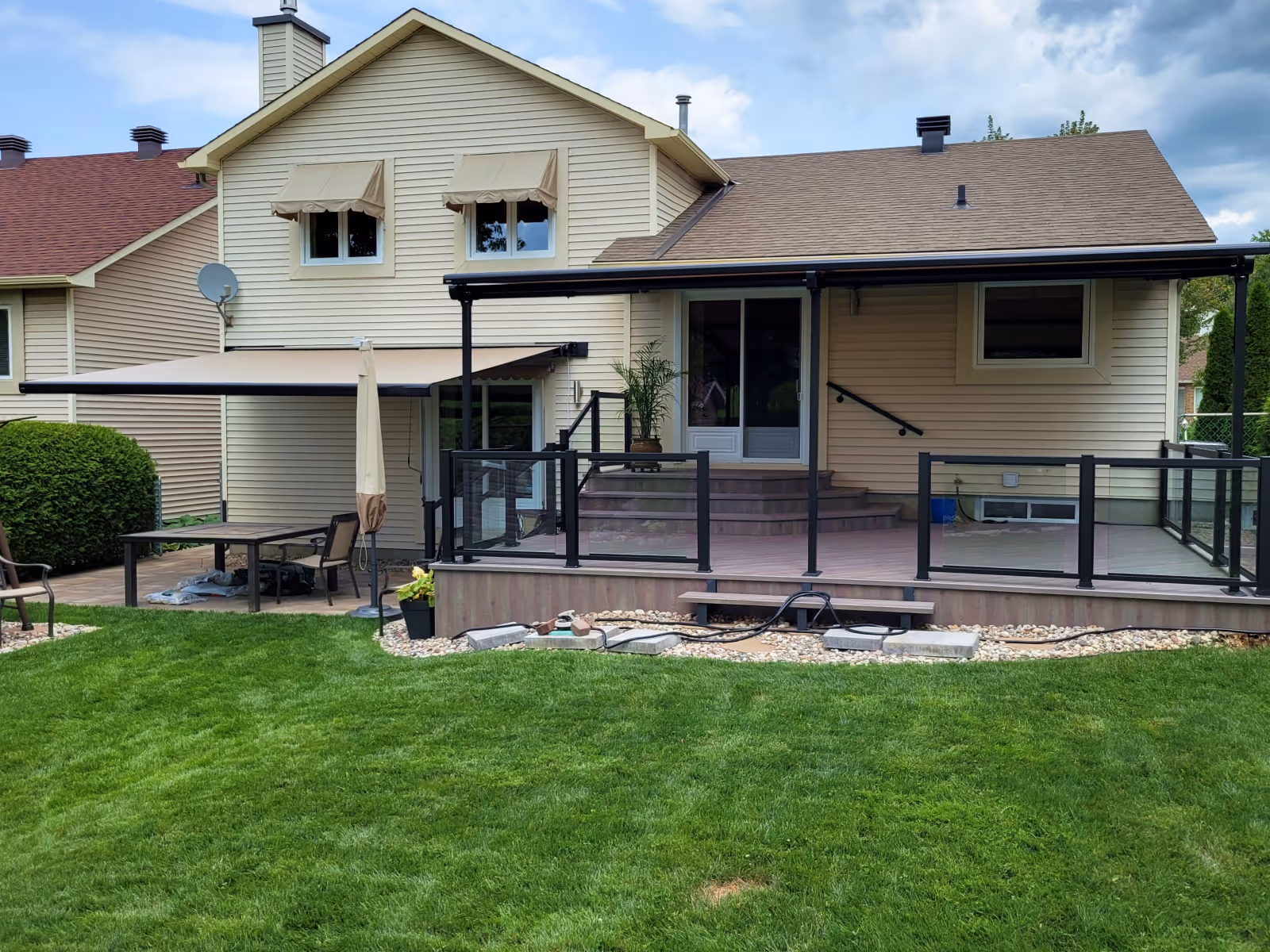 A wood deck with glass railings is covered by a Pergolino awning, a stone patio is shaded by a retractable awning, and two second-storey windows have window awnings. All have beige fabric.