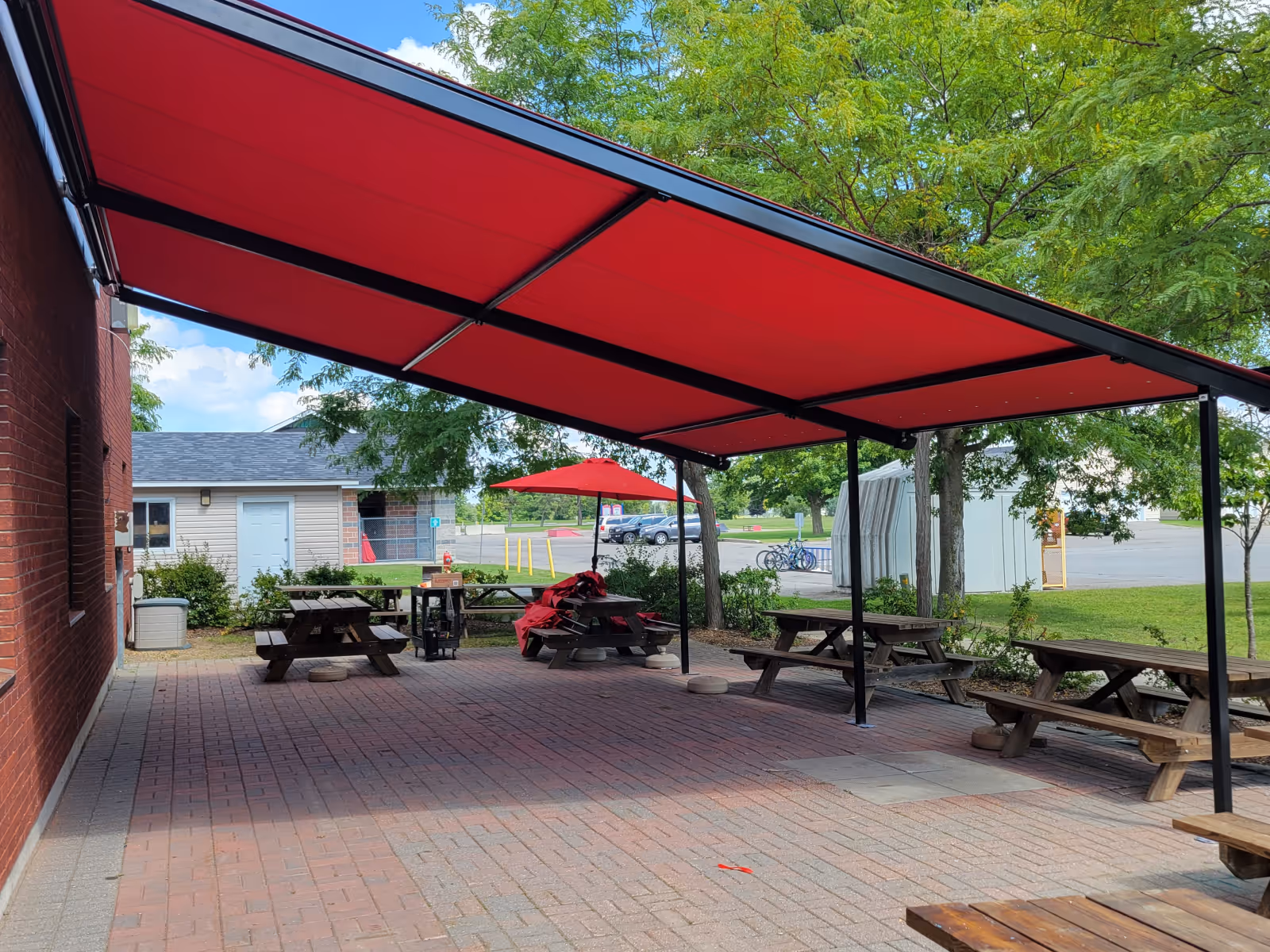 A large Pergolino awning with red fabric shades a stone patio with wood picnic tables next to a brick building.