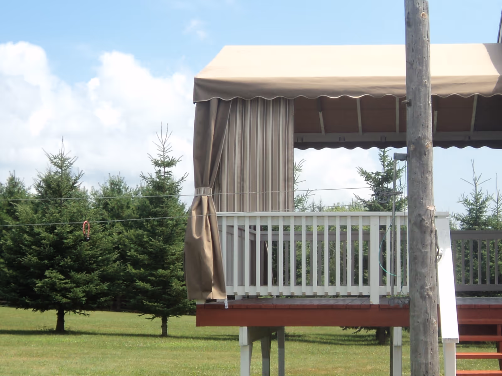 An a-frame fixed awning with a solid beige fabric cover and a coordinating beige stripe track curtain covers an elevated wood deck with a white railing.