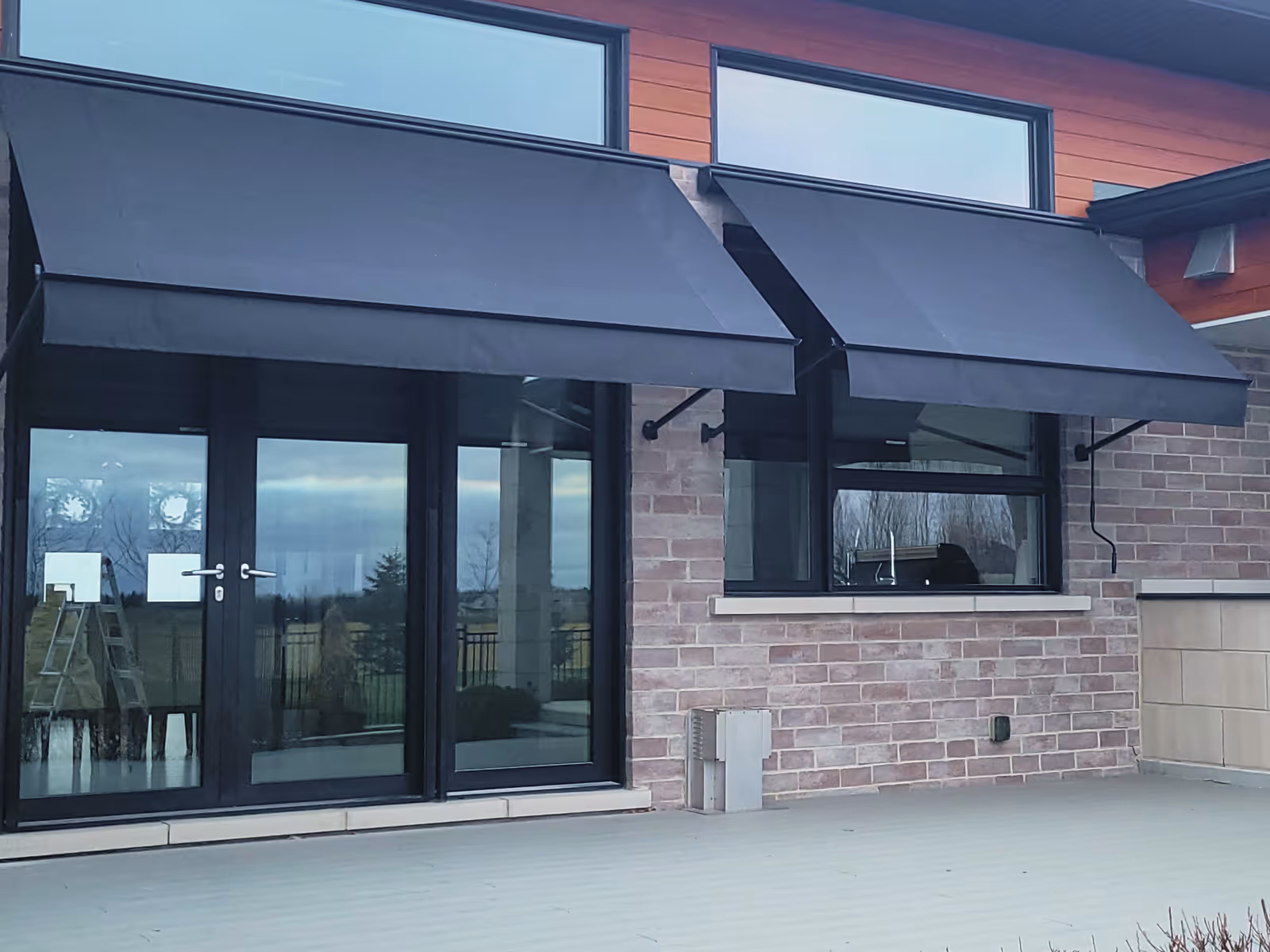 A glass door and a window are shaded by a pair of black awnings that sit below transom windows on a commercial building.