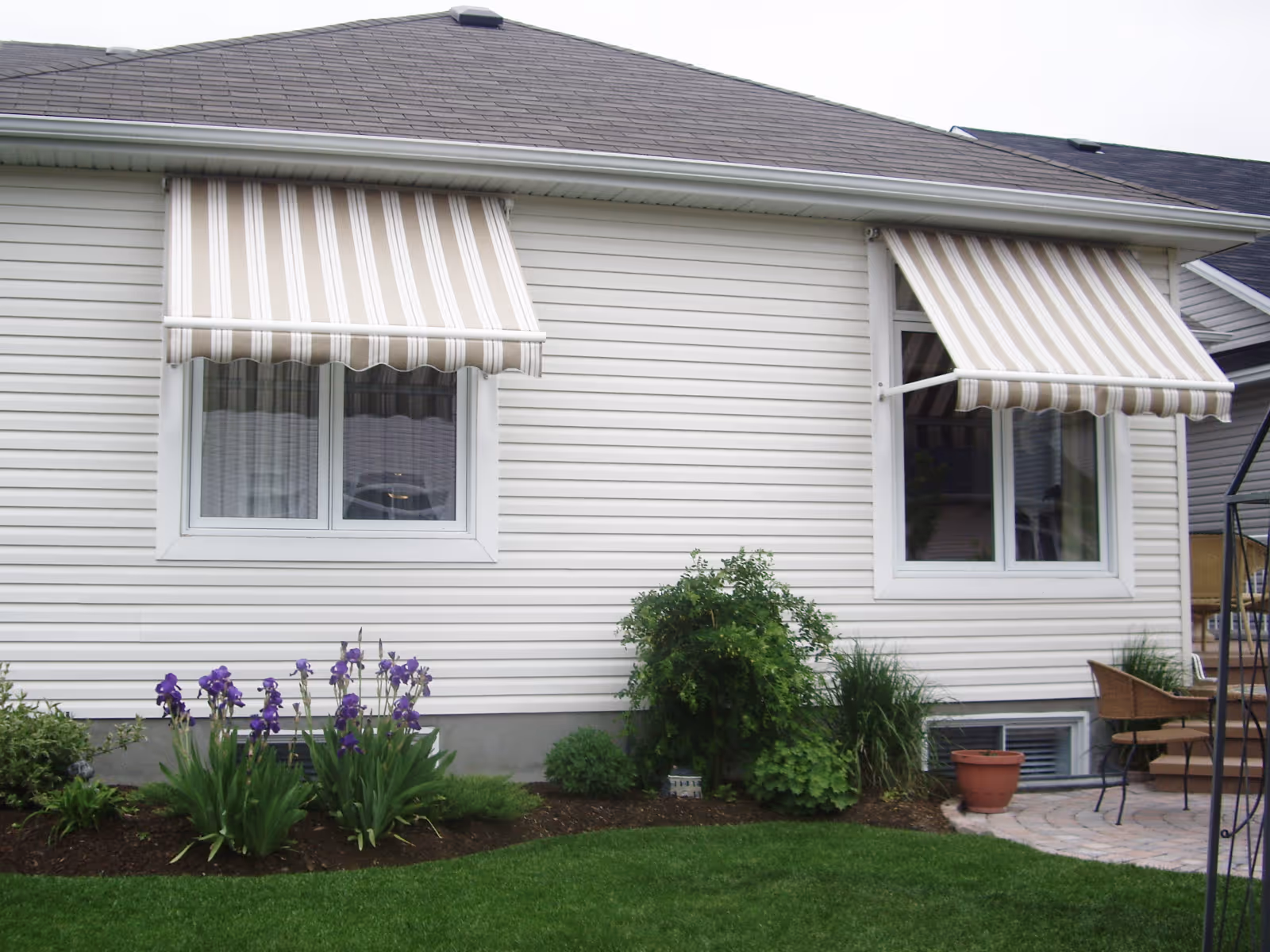A pair of beige and white awnings shade windows above a flowerbed and a stone patio.
