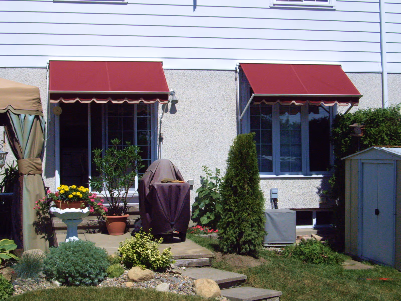 A pair of burgundy retractable window awnings on a house with grey stucco and white siding.
