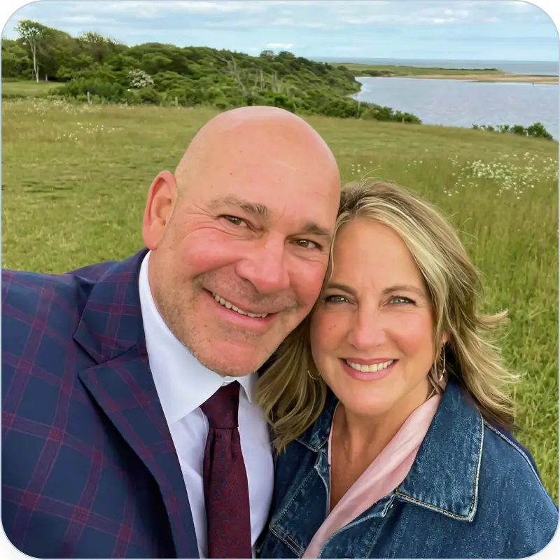 Smiling man in a checkered suit and woman in a denim jacket posing together outdoors with a grassy field, trees, and water in the background.