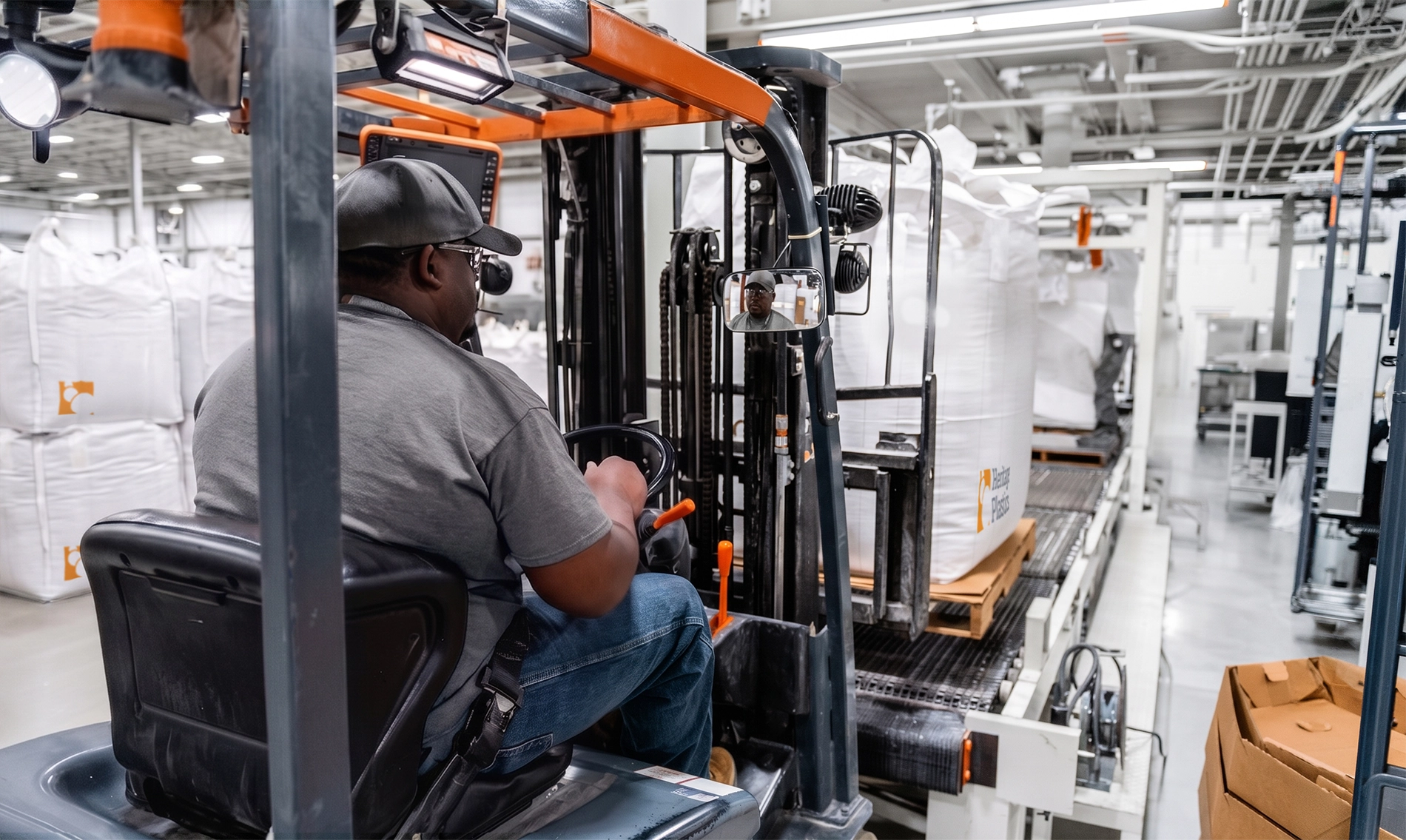 Worker operating forklift in warehouse lifting large white bags of plastic resin pellets labeled Heritage Plastics.