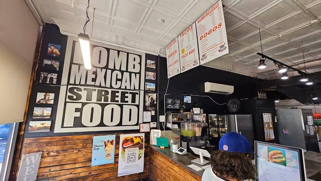 Counter area of a casual restaurant with “BOMB MEXICAN STREET FOOD” sign on the wall, menu boards above, drink dispensers, and a person standing at the counter.
