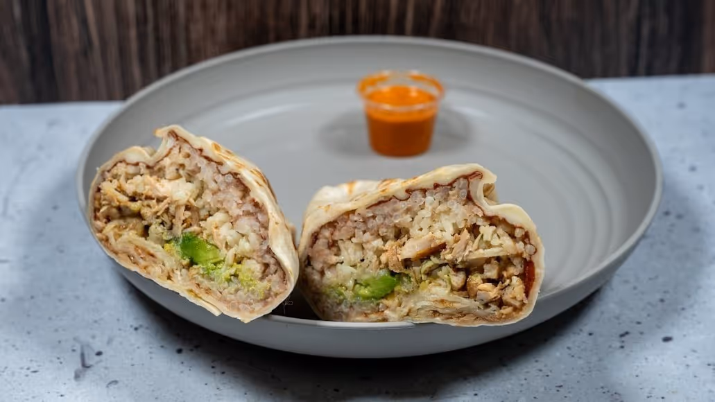 A sliced burrito revealing rice, chicken, and avocado inside rests on a gray plate. A small cup of orange sauce is in the background, creating a savory and appetizing scene.
