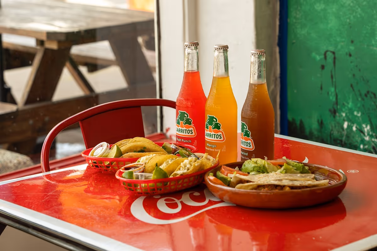 A colorful spread of Mexican food and drinks on a red Coca-Cola table. Three Jarritos bottles stand behind tacos and lime wedges in red baskets.