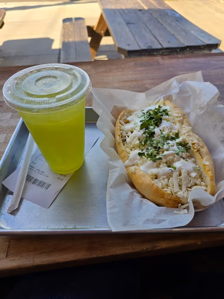 A hot dog topped with cheese, cilantro, and white sauce on a tray next to a vibrant green drink in a plastic cup. Background shows wooden picnic tables.