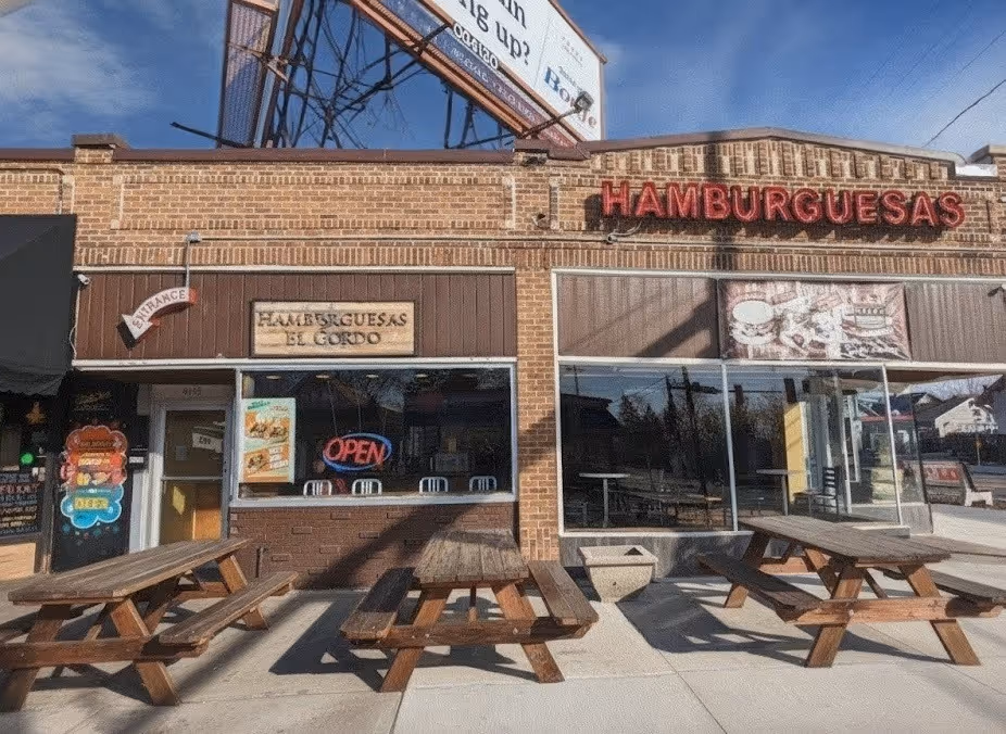 Brick restaurant facade with "Hamburguesas" sign and "Open" neon light. Three wooden picnic tables sit outside under a clear blue sky. Cozy, inviting vibe.