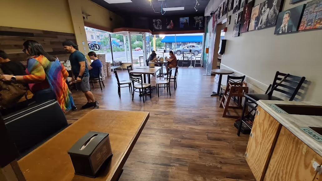 A cozy cafe interior with wooden floors, empty tables, and chairs. People sit by large windows, and colorful artwork decorates the cream walls.