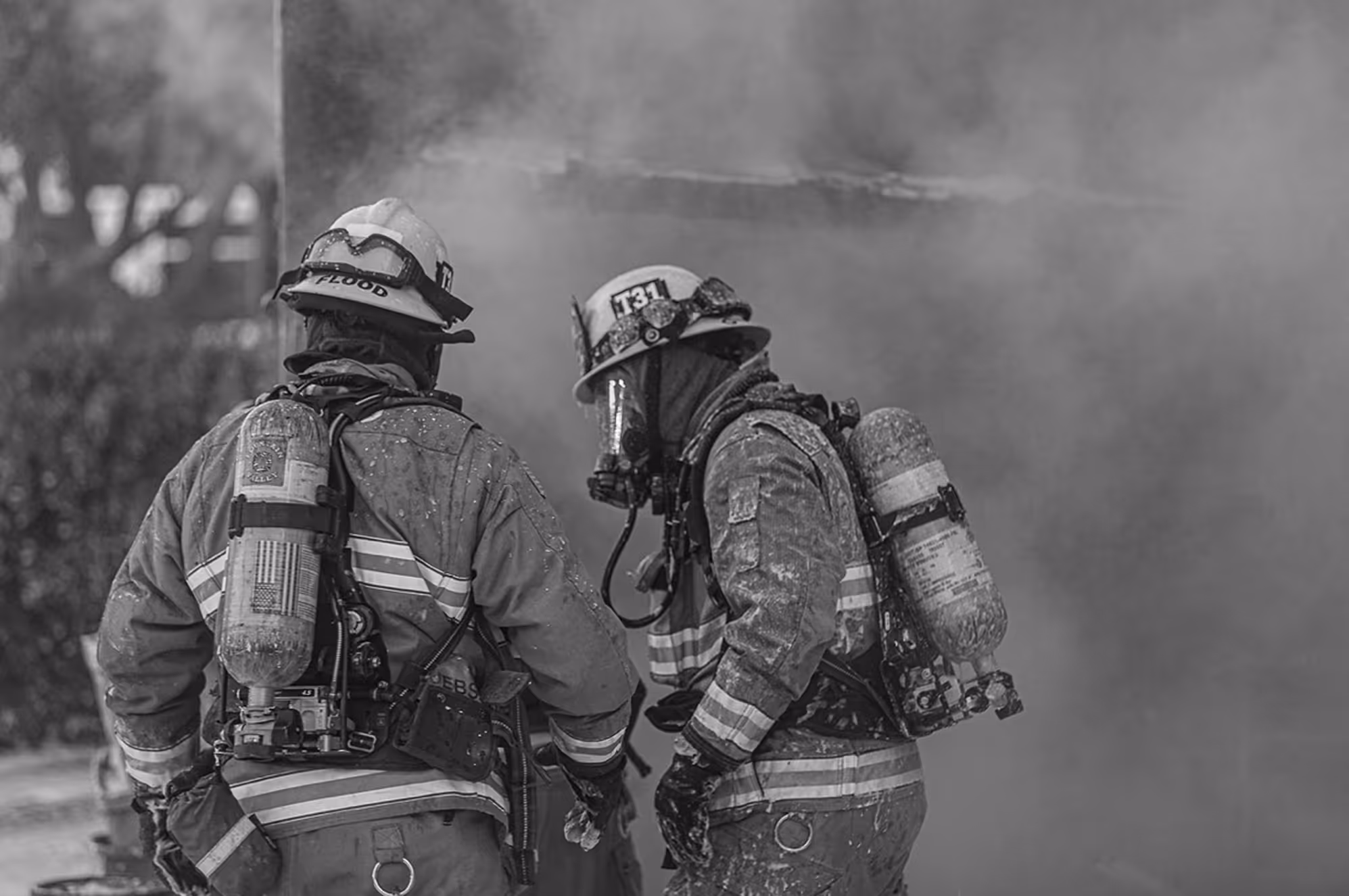 Two firefighters in full gear with breathing apparatus facing away amid thick smoke.