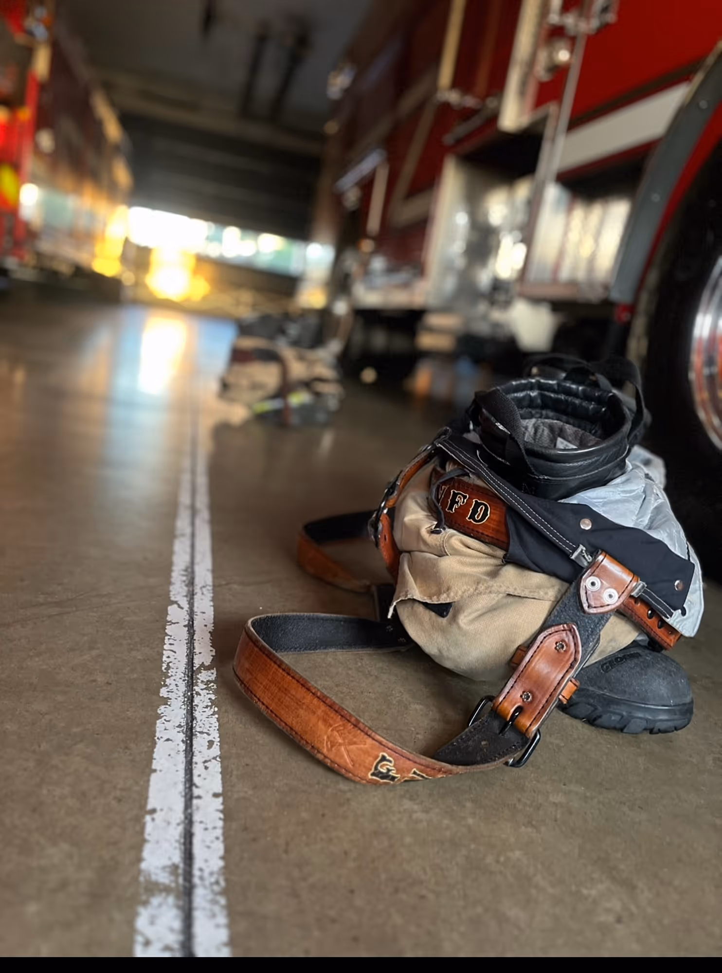 Firefighter gear including boots and jacket laid on the floor of a fire station near a fire truck.