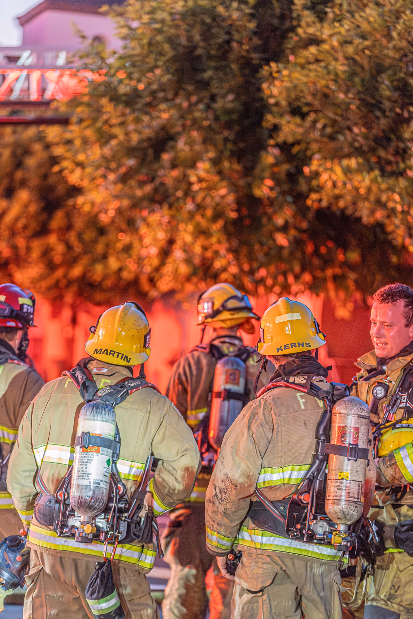 Firefighters in full gear with oxygen tanks on their backs standing outside during a fire emergency at dusk.