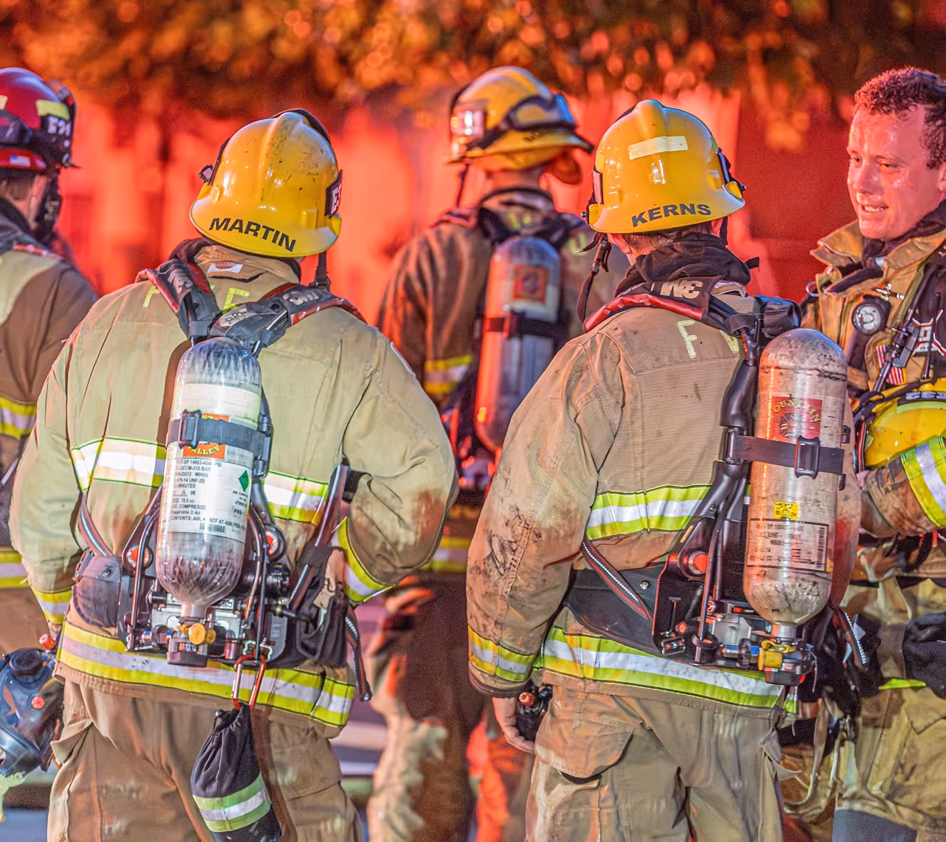 Group of firefighters in full gear, including helmets and oxygen tanks, facing away with a glowing orange background.