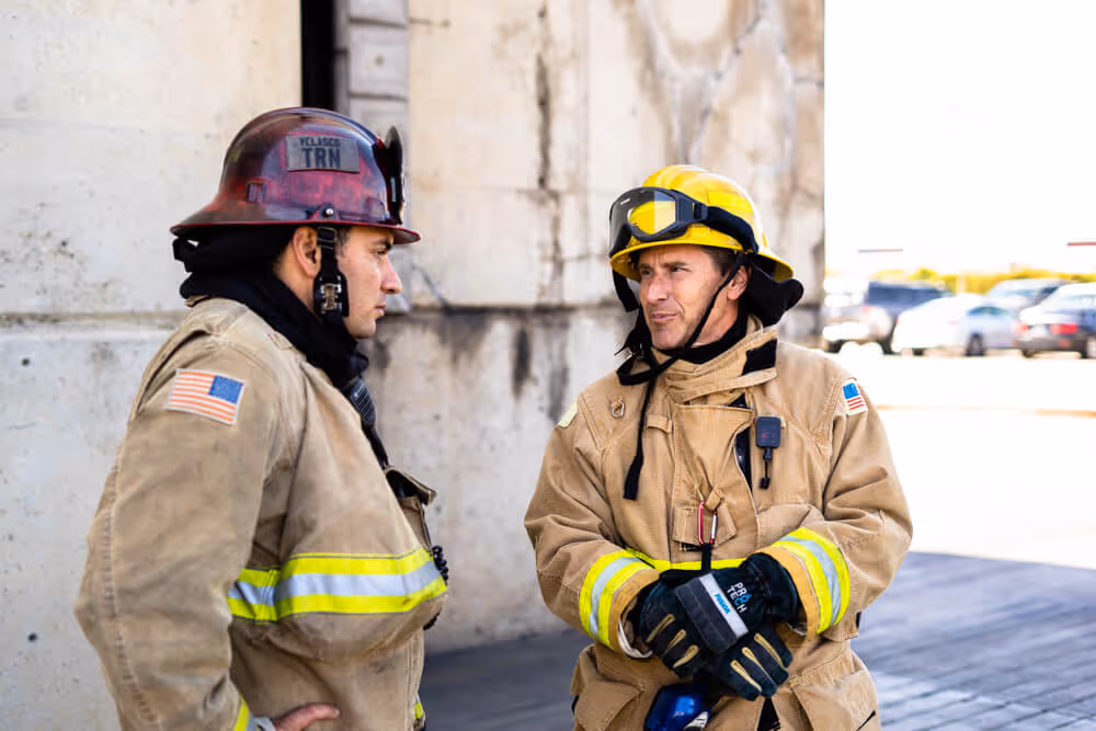 Two firefighters in beige uniforms and helmets having a serious conversation outside near a concrete wall.