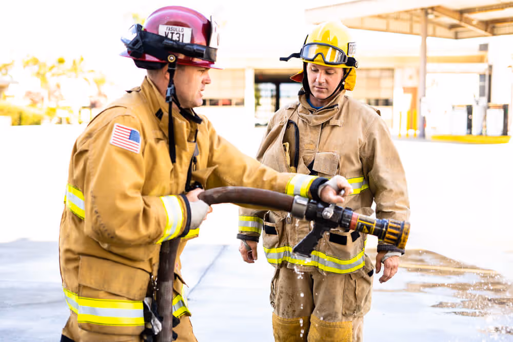 Two firefighters in full gear holding a fire hose outdoors with water spraying.